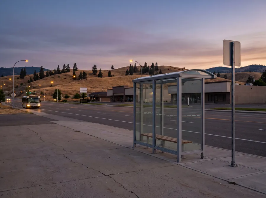 Generated picture: an empty bus shelter on a wide road at dusk, a bus with its lights on further down the road and grassy hills beyond.